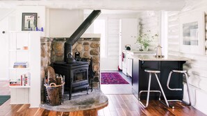 A cozy room with a stone fireplace, wooden flooring, a small kitchen area with a bar stool, and a bookshelf.