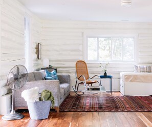 A cozy room with a wooden floor, a grey sofa with a throw blanket, a wicker chair, a small table with a vase, and a fan.