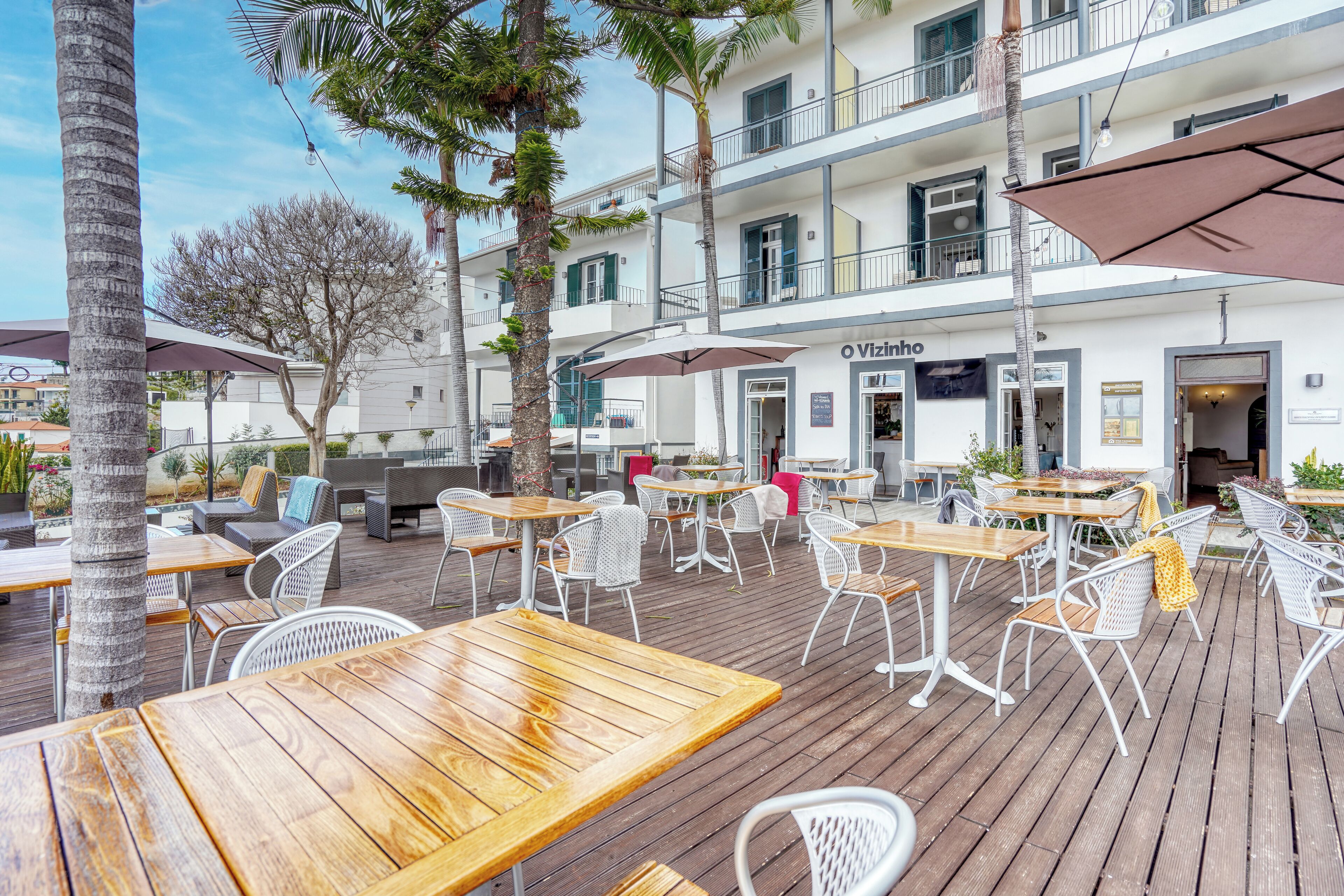 An outdoor seating area with wooden tables and white chairs, a building with balconies, and a sign that reads 'O Vizio'.