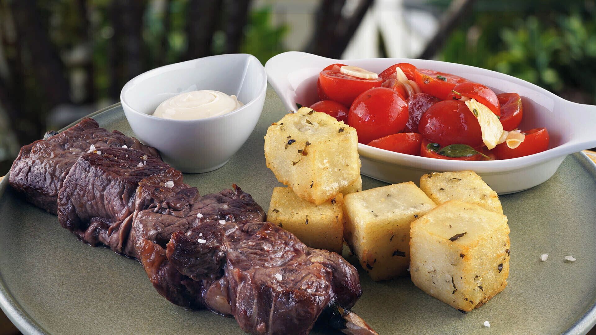 A plate with grilled meat, roasted potatoes, and a bowl of tomatoes.