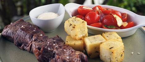 Une assiette avec de la viande grillée, des pommes de terre rôties et un bol de tomates.