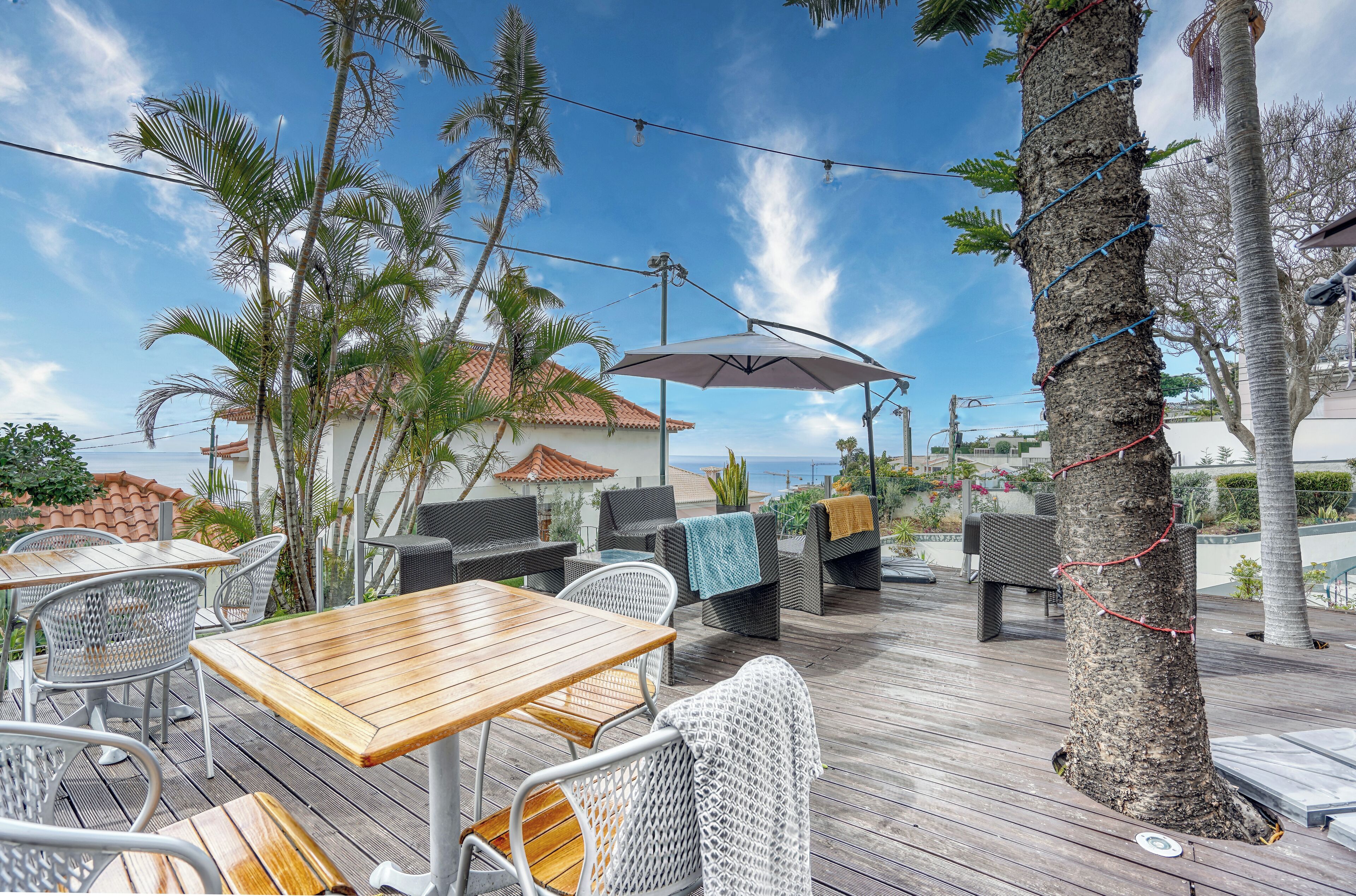 An outdoor patio with wooden tables and chairs, surrounded by palm trees and a view of the sea.