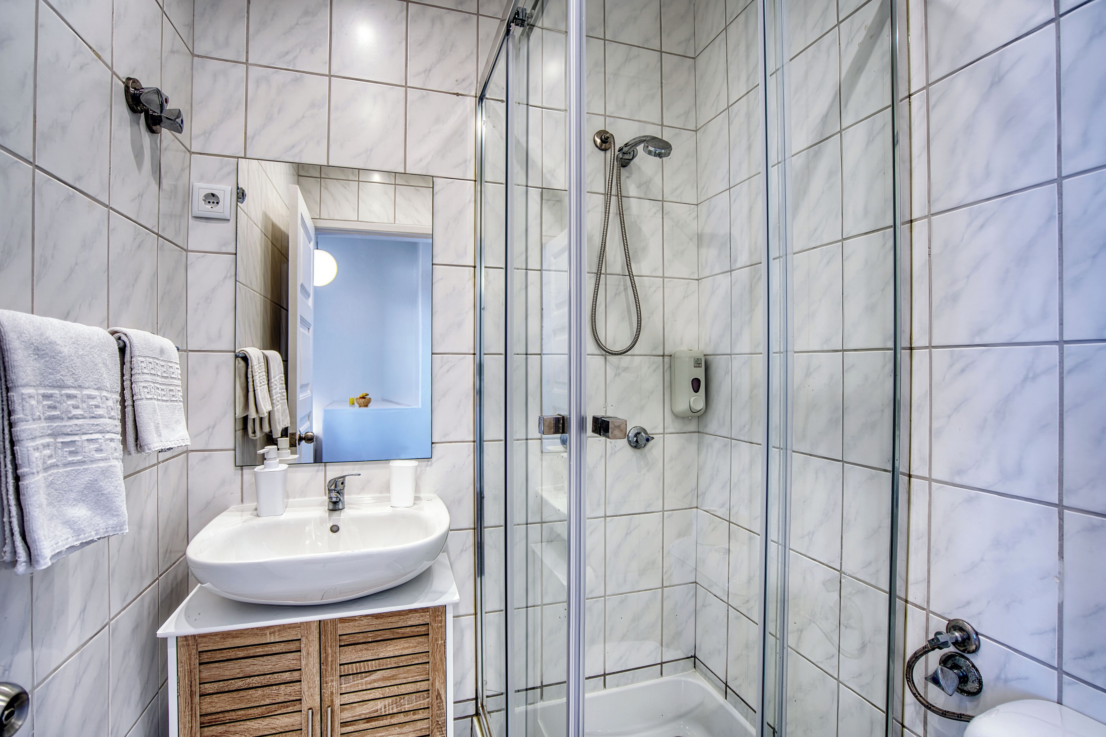 Une salle de bain avec un lavabo blanc, un meuble-lavabo en bois, un miroir, une douche avec une porte en verre et des murs carrelés.