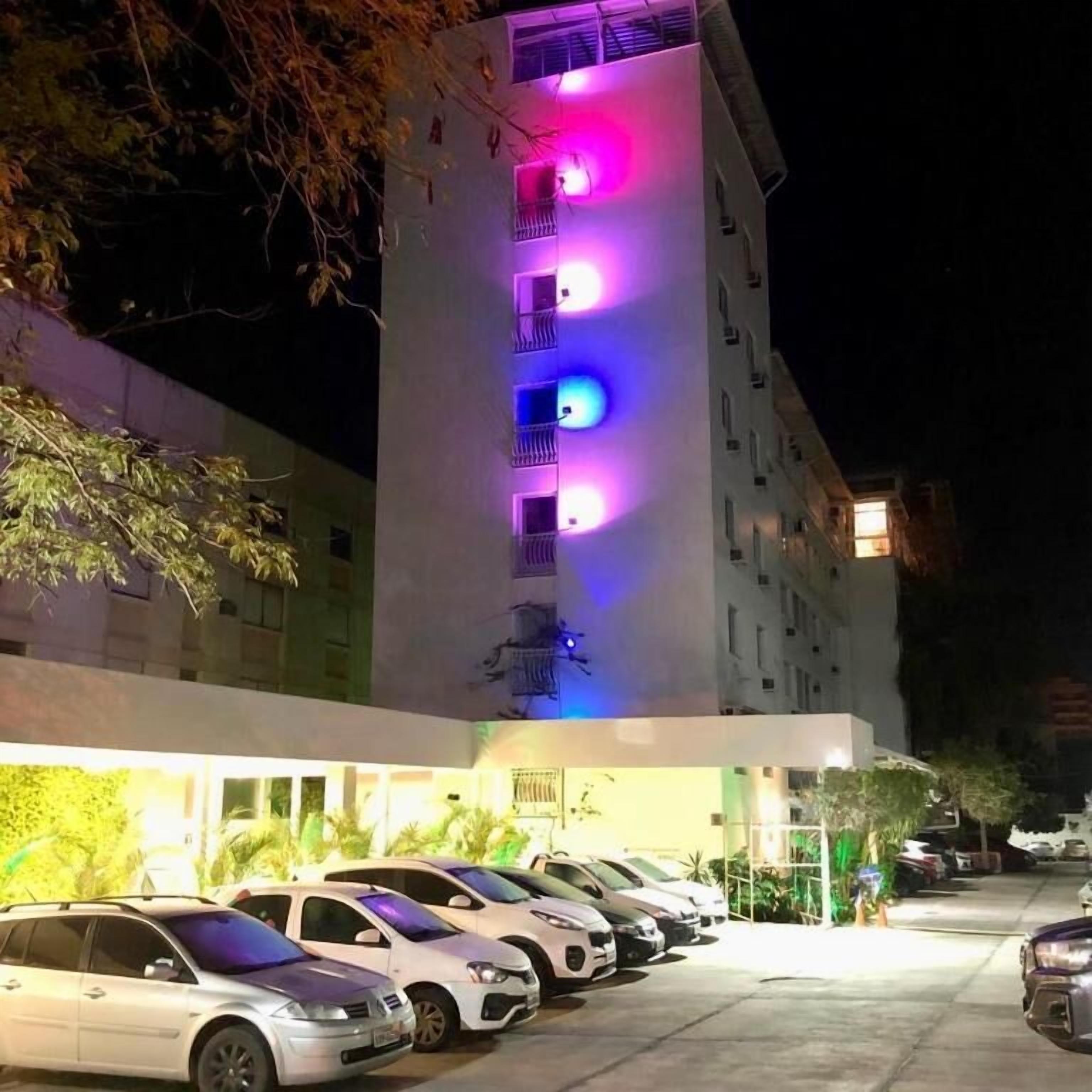 A hotel with a row of parked cars in front and colorful lights on the building.