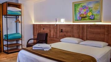 Quarto de hotel com uma cama grande, cabeceira de madeira, uma cadeira, prateleira com roupas dobradas e um quadro na parede.