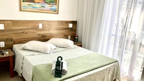 A hotel room with a wooden headboard, a bed with white linens, a green throw blanket, a towel with a green tag, a nightstand with a phone, and a painting on the wall. - HOTEL APPEL (Santa Maria)