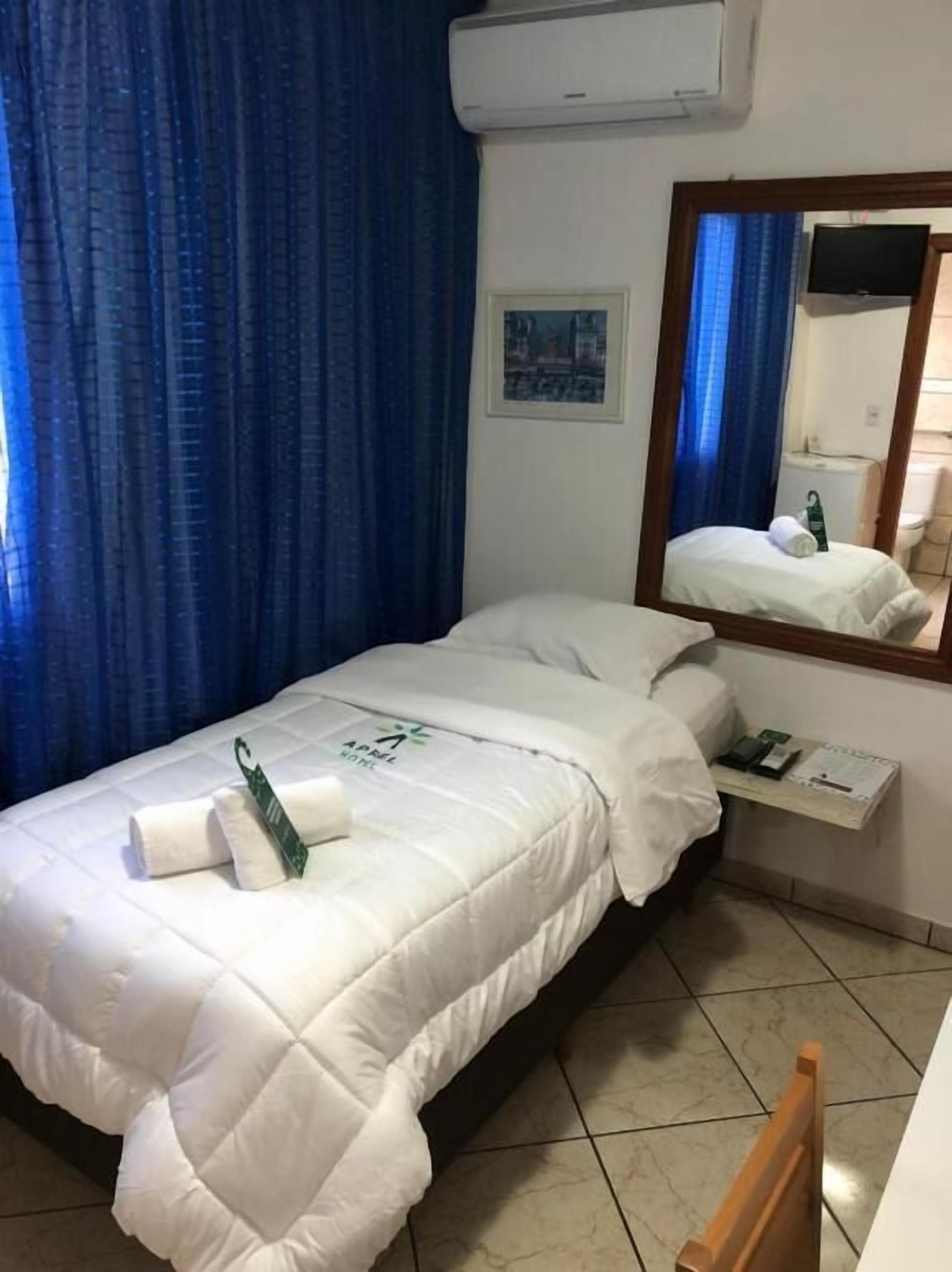 A hotel room with a bed, a small bedside table with a lamp, a mirror, and a window with blue curtains.