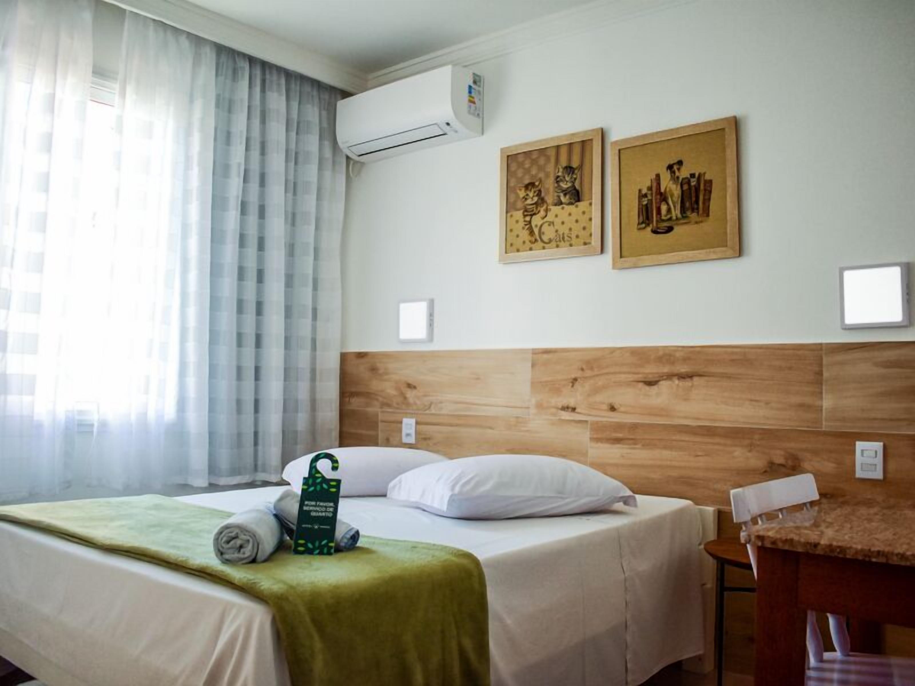 A hotel room with a wooden headboard, a bed with white linens, a green blanket, and a small table with a chair. There are two framed artworks on the wall and an air conditioning unit above the bed.