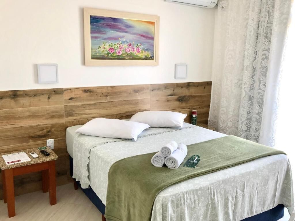 Quarto de hotel com cabeceira de madeira, cama com roupa de cama branca, manta verde e um quadro na parede.
