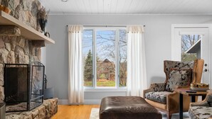 A cozy living room with a stone fireplace, a large window with curtains, a leather sofa, and a patterned armchair. - Wooded & Quiet, but Downtown. Spacious Home w/ Smokeless Fire Pit, Barrel Sauna (Bellaire)
