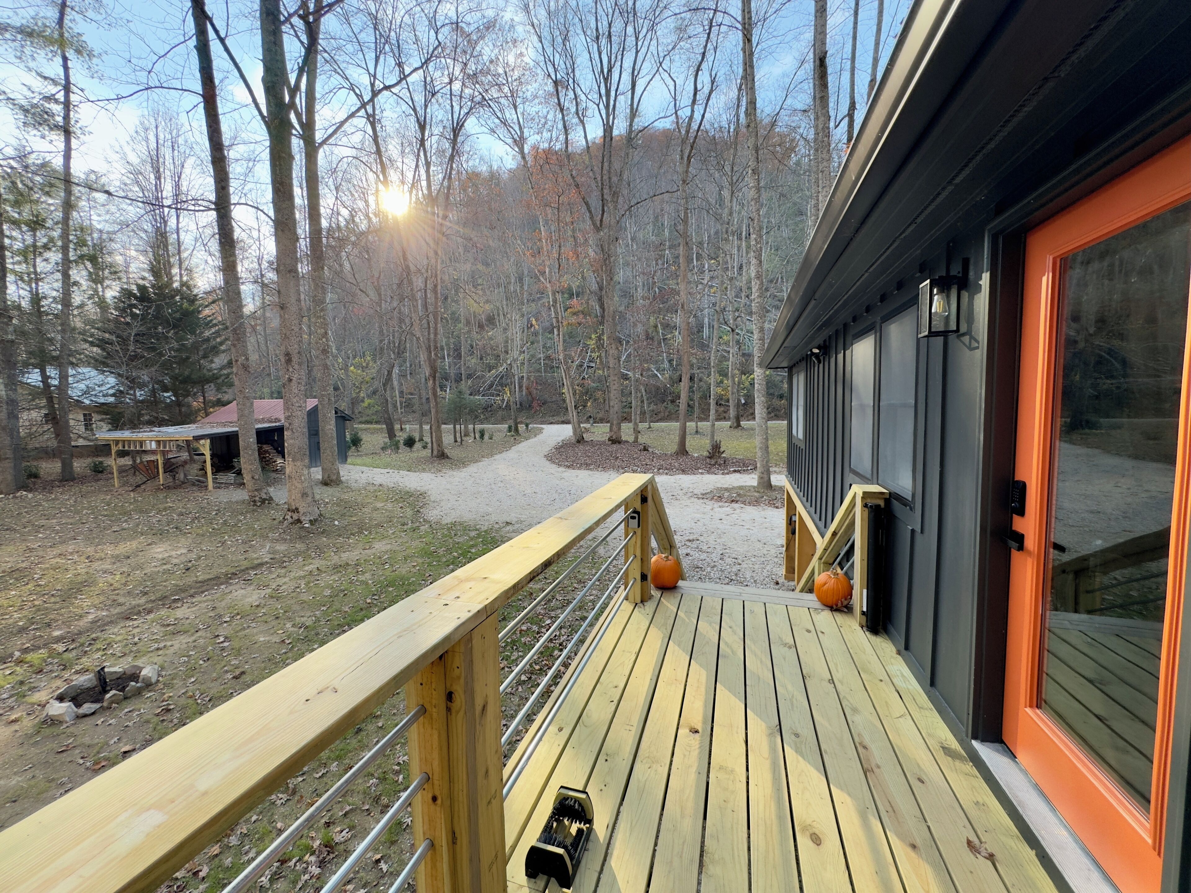A wooden deck with a railing, a pathway leading to a building with a black exterior and orange trim, and a forested background.