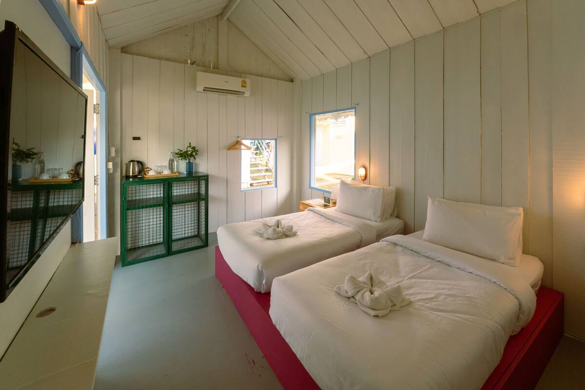 A room with two beds, a green cabinet, a window with a view, and an air conditioning unit.