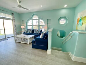 Ein geräumiges Wohnzimmer mit blauem Sofa, einem weißen Couchtisch und großen Fenstern.