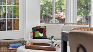 A cozy living room with a white sofa, a colorful throw pillow, a wooden tray with a plant, and a window with a view of greenery.