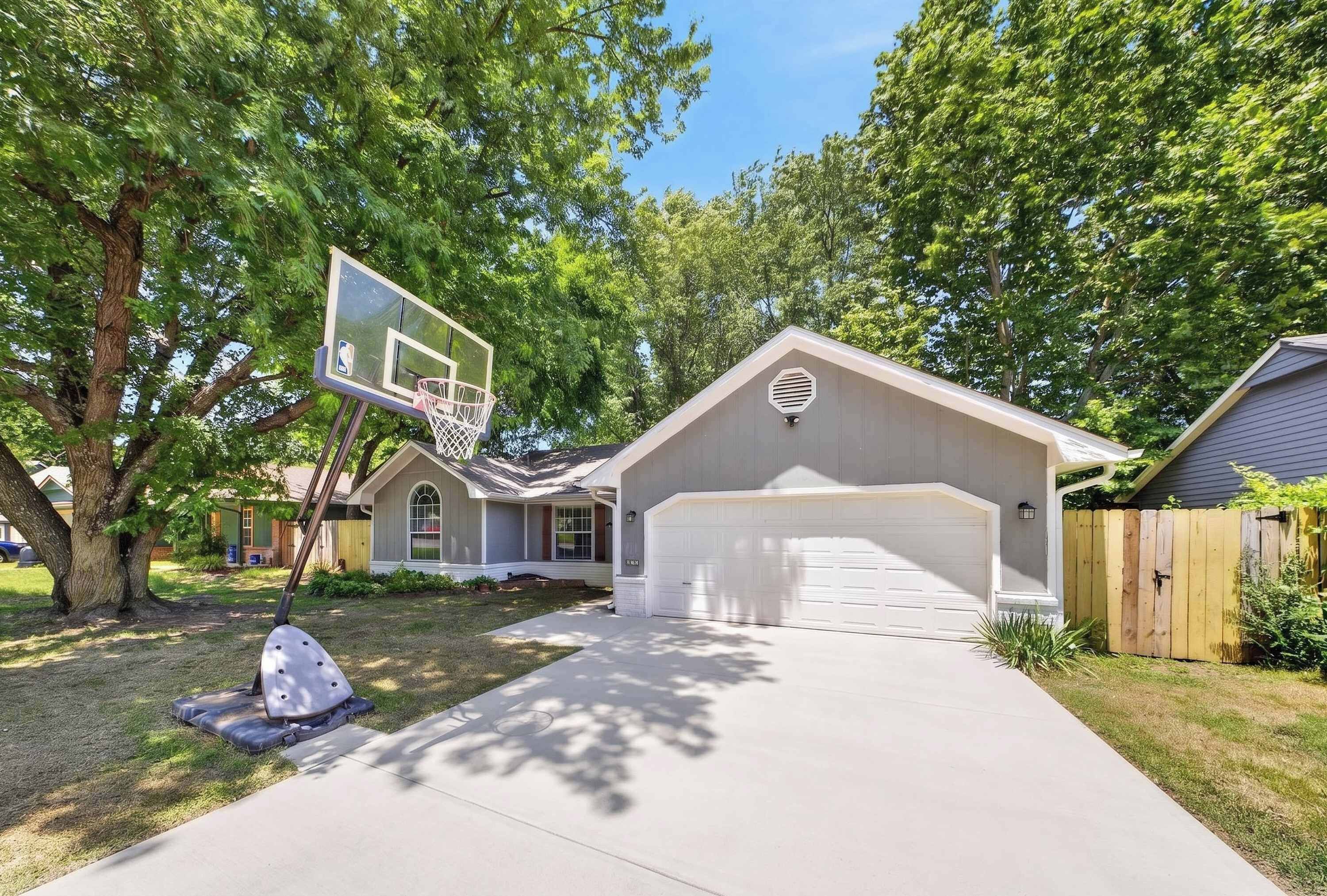 A residential house with a garage, a basketball hoop, and a tree in the yard.