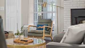 A living room with a round coffee table, a green armchair with a leopard print pillow, a floor lamp, and a fireplace.