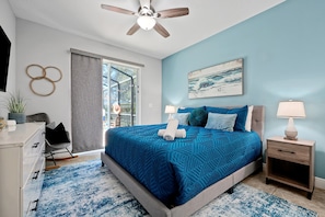 Une chambre à coucher comprenant un lit, deux tables de chevet, un ventilateur de plafond, une fenêtre avec des rideaux, une œuvre d’art accrochée au mur et une petite plante.
