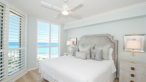 A bedroom with a large bed, a ceiling fan, a nightstand with a lamp, and a view of the ocean through the window. - Serene Relaxation with Semi-Private Elevator at St. Maarten (Destin)
