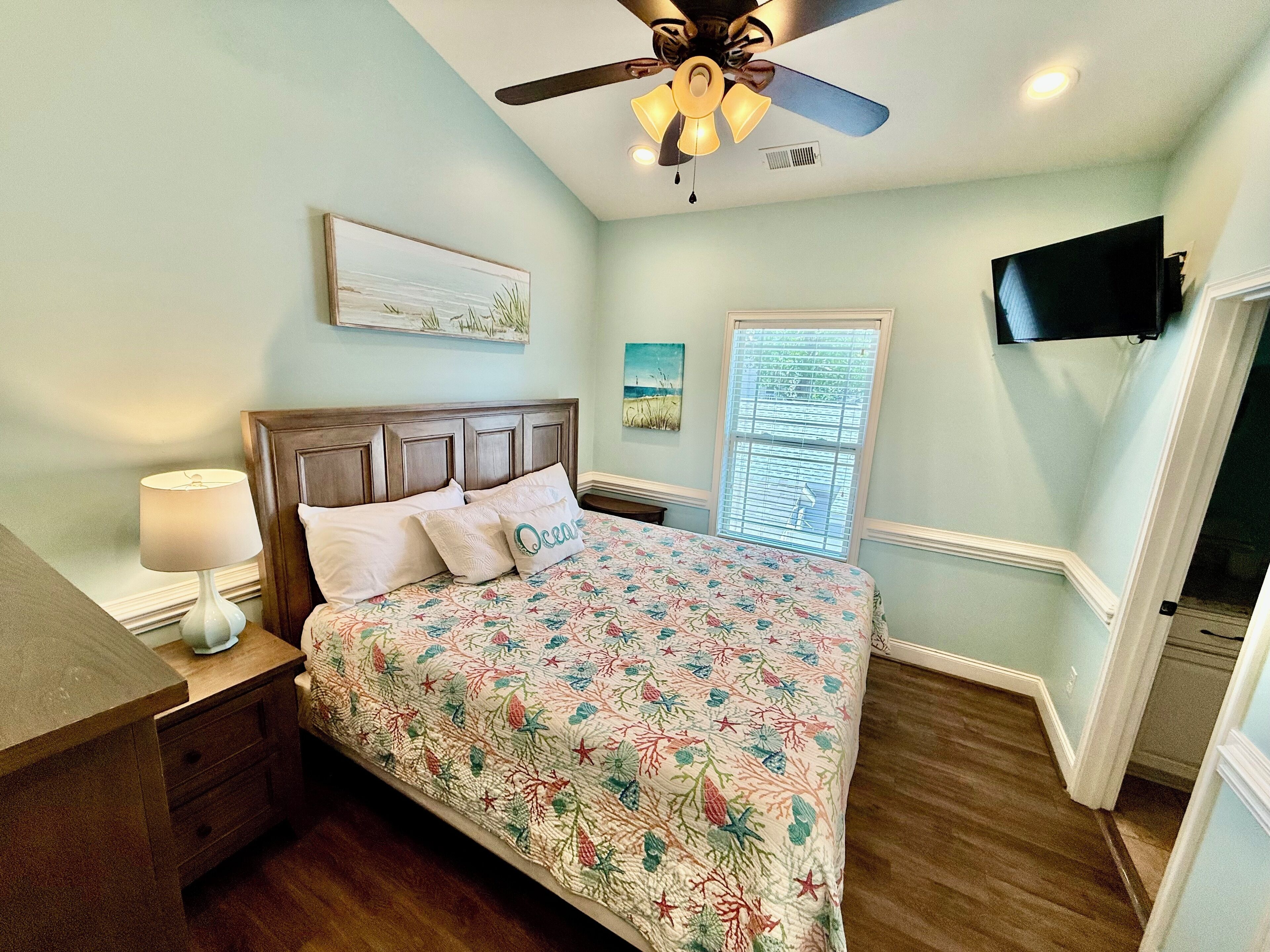 A bedroom with a bed, nightstand, lamp, ceiling fan, artwork, window, and a TV mounted on the wall.