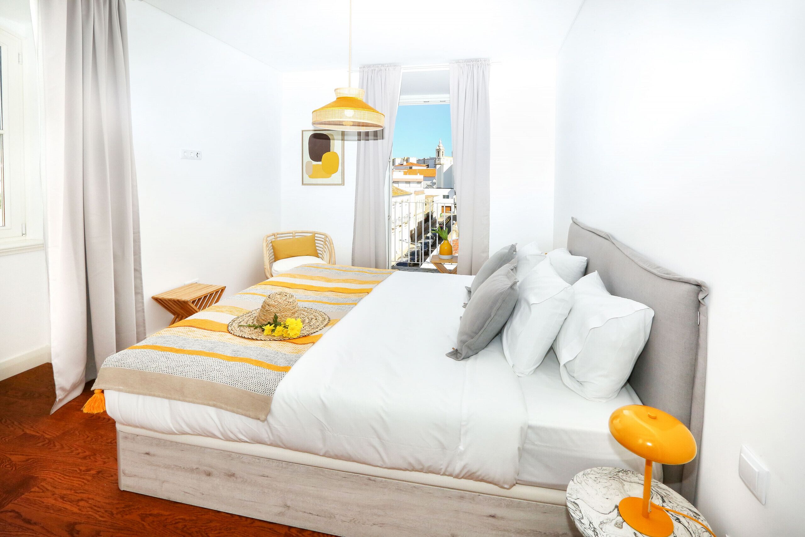 A bedroom with a bed, a bedside table, a lamp, and a view of buildings through a window.