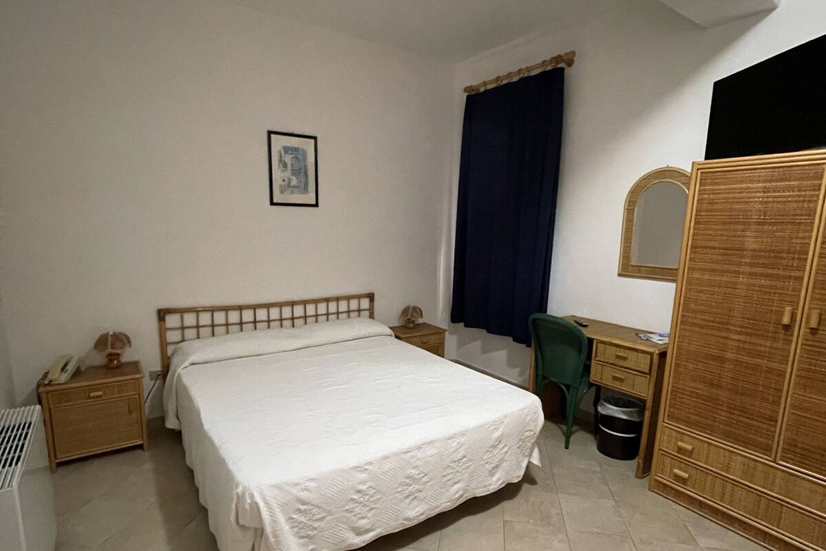 A bedroom with a bed, a desk, a chair, a wardrobe, a mirror, a nightstand, and a radiator.
