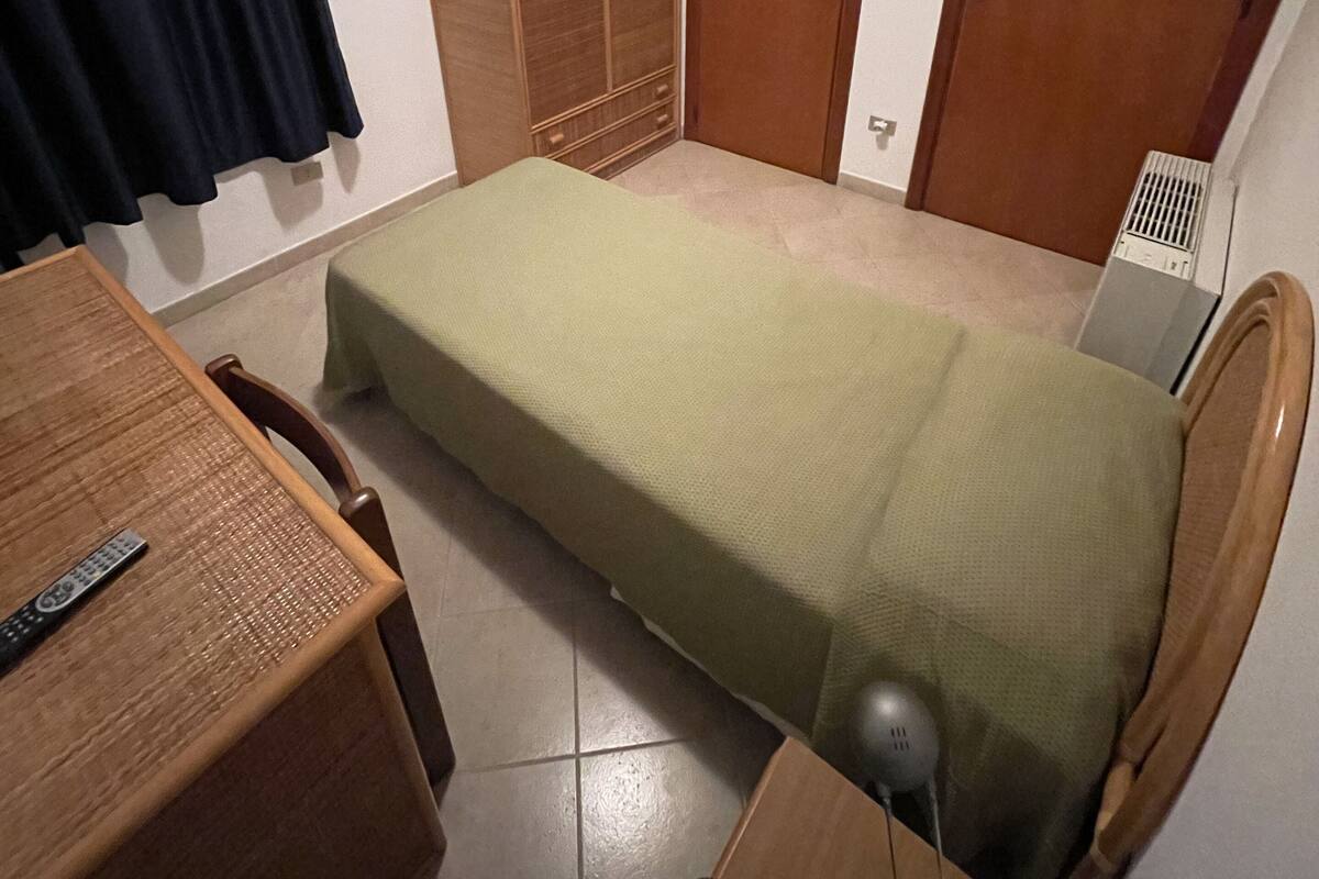 A small, neatly arranged room with a single bed, a wooden wardrobe, a desk with a chair, and a television on the wall.