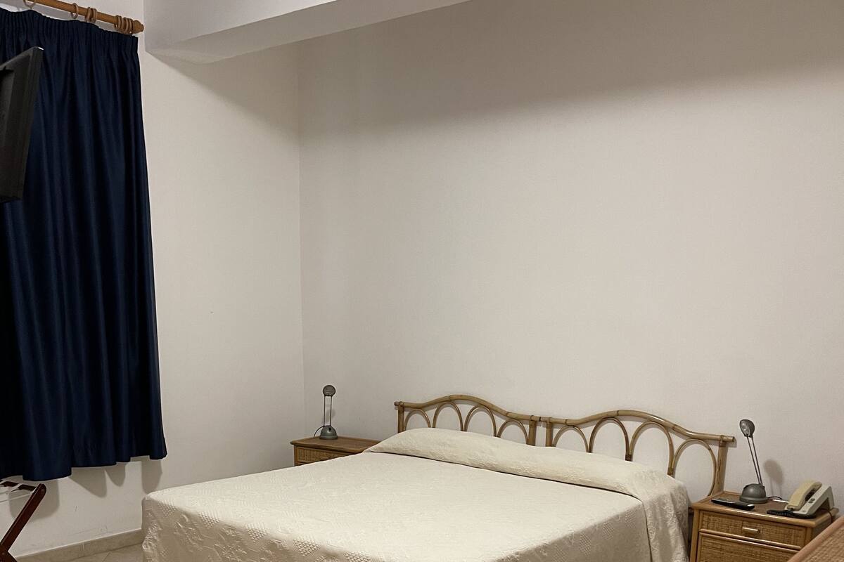 A bedroom with a bed, a nightstand, a lamp, a telephone, and a curtain.