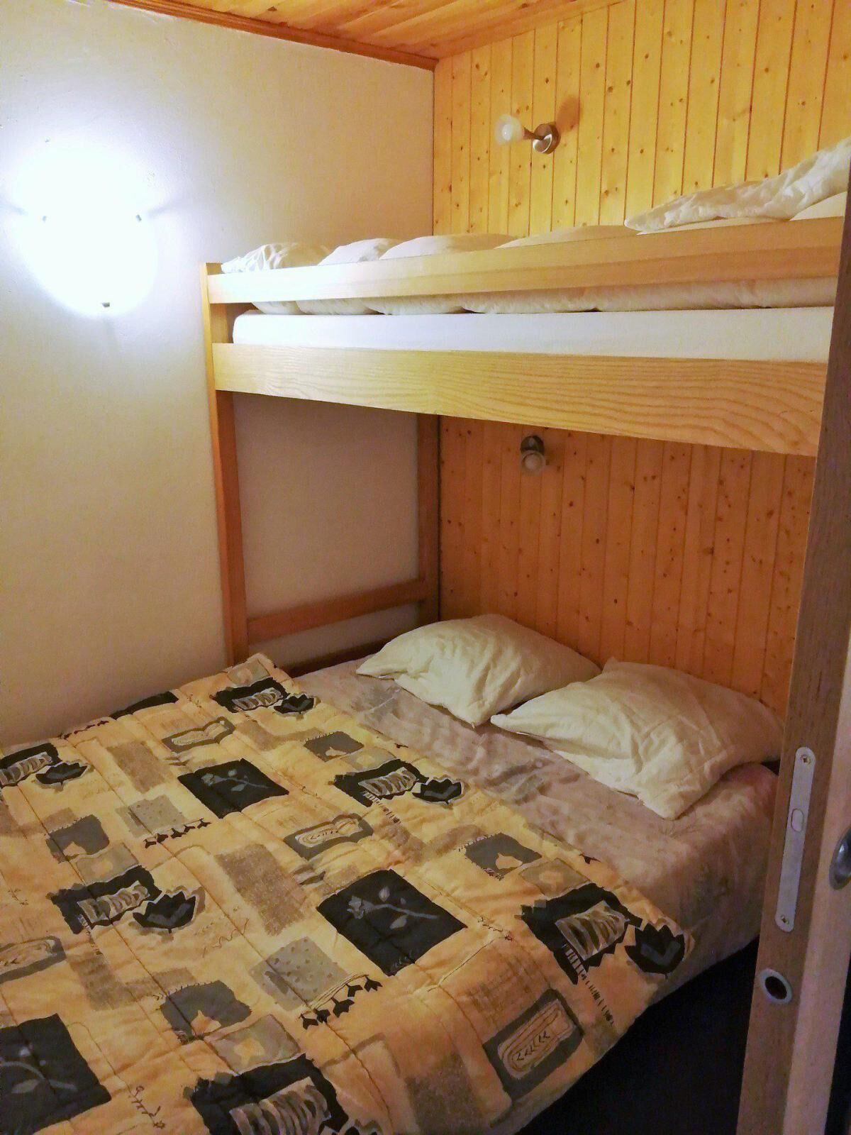 Een slaapkamer met stapelbed, houten plafond en muren, een gewatteerde sprei en een lamp aan de muur.