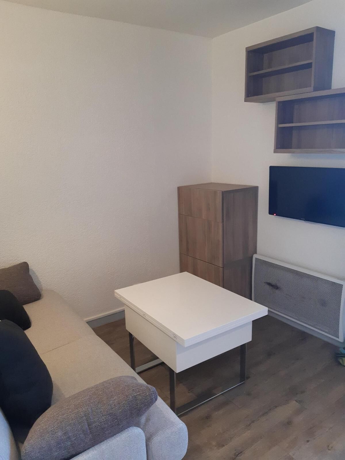 A modern living room with a white sofa, a small white table, and a wooden cabinet. There are built-in shelves above the sofa and a flat-screen TV mounted on the wall.