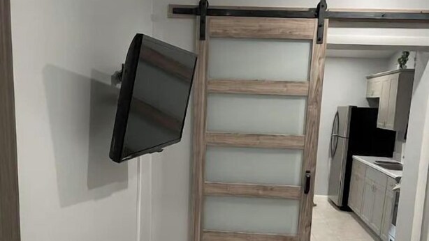 Una habitación con puerta corredera, televisor montado, zona de cocina al fondo y suelo de baldosas.