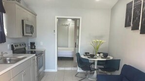 Una cocina compacta con microondas, cafetera y lavabo. Hay una zona de comedor con mesa, sillas y un jarrón de flores. Un sofá azul está en primer plano.