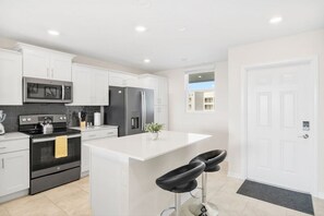 A modern kitchen with white cabinetry, stainless steel appliances, a central island with bar stools, and a view of the outside through a window.