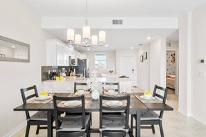 A modern dining area with a rectangular table set for six, surrounded by black chairs. A chandelier hangs above the table, and a kitchen is visible in the background.
