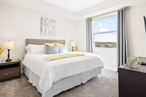 A modern hotel room with a large bed, a nightstand, a lamp, and a view of the outdoors through a window.