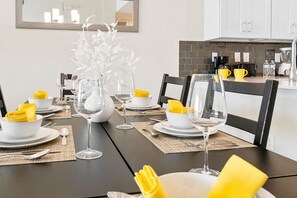 A dining table set with white plates, yellow napkins, and clear wine glasses, with a kitchen in the background.