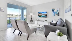 A modern living room with a sofa, armchairs, a coffee table, and a TV. There is a large window with a view of a body of water and a building.