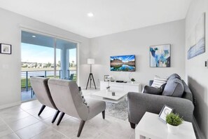 A modern living room with a sofa, armchairs, a coffee table, and a TV. There is a large window with a view of a body of water and a building.