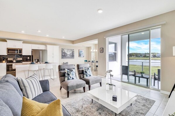 A modern living room with a grey sofa, two armchairs, and a coffee table. There is a kitchen area with white cabinets and a dining area with a table and chairs. A large sliding glass door leads to a balcony with a view of a green landscape.