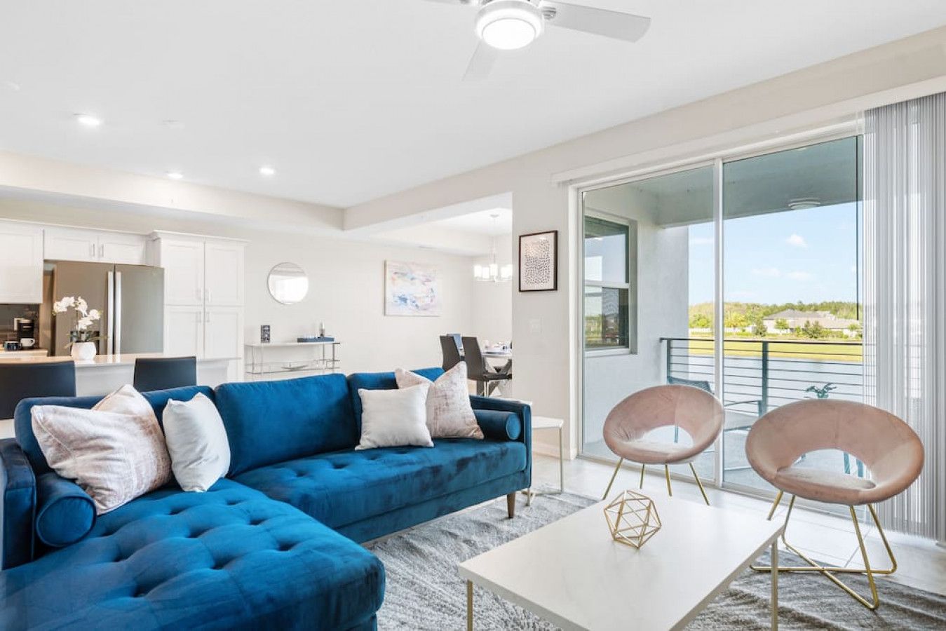 A modern living room with a blue sectional sofa, a white coffee table, and two chairs. There is a kitchen in the background and a balcony with a view.