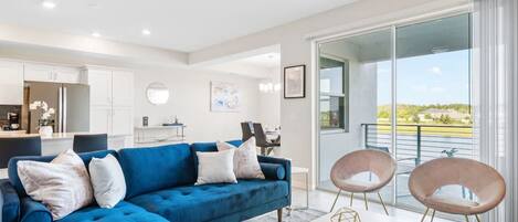 A modern living room with a blue sectional sofa, a white coffee table, and two chairs. There is a kitchen in the background and a balcony with a view.