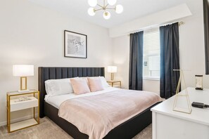 A modern bedroom with a black headboard, two bedside tables with lamps, a large bed with a pink blanket, a framed artwork above the bed, and a window with dark curtains.