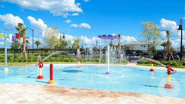 A swimming pool with multiple water features, including fountains and water jets, surrounded by a tiled deck and palm trees.