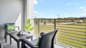Un balcon donnant sur un espace verdoyant et une route. On y trouve une table et deux chaises avec des coussins.