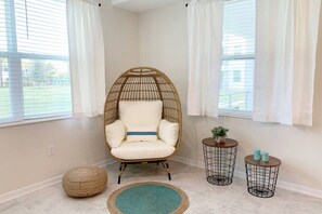 A room with a hanging chair, two side tables, a round rug, and a woven pouf.