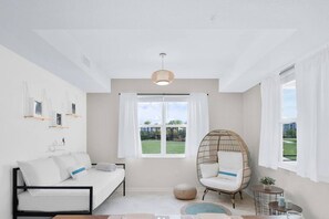 A bright, minimalist living room with a white sofa, a wicker chair, a round table, and a large window offering a view of a green landscape.