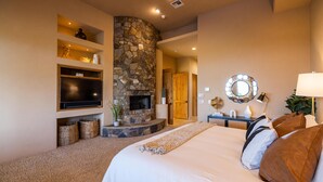 A bedroom with a stone fireplace, a flat-screen TV, a large bed with multiple pillows, and a bedside table with a lamp. - Luxury Hideout with Infinity Pool & Spa (Scottsdale)