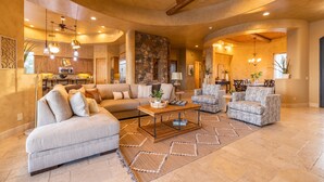 A spacious living room with a sectional sofa, a coffee table, and patterned armchairs. A stone fireplace and kitchen area are visible in the background. - Luxury Hideout with Infinity Pool & Spa (Scottsdale)