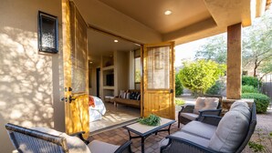 An open patio with wicker furniture, a wooden door leading to an interior space, and a view of greenery outside. - Luxury Hideout with Infinity Pool & Spa (Scottsdale)