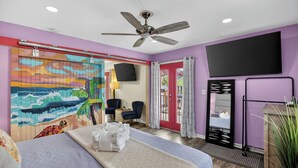 Ein Hotelzimmer mit Bett, Deckenventilator, an der Wand montiertem Fernseher und einer Schiebetür mit Strandgemälde.