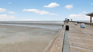 Ein hölzerner Steg führt zu Strandhäusern, mit einem Schild, das den Gezeitenplan anzeigt.